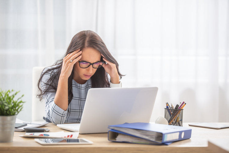 A woman suffering from migraine sits at her laptop and holds her head – an illustration of headache and seeking relief with CBD oil for migraines.