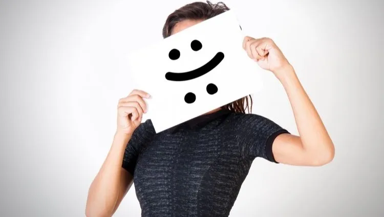 A woman holds a piece of paper with a smiley face in front of her face as a symbol of changing moods and emotions in connection with the topic of bipolar disorder and the possibility of support using CBD oil