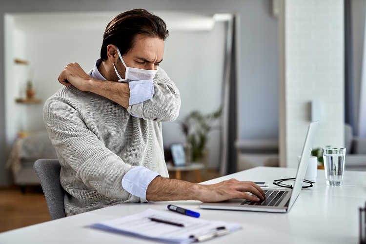 A young man sits at a computer wearing a protective mask and coughing into his elbow