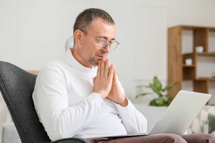 Man wearing glasses sitting in an armchair, focusing on his laptop with hands clasped near his face.