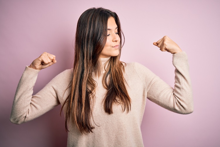 A woman with long brown hair, wearing a beige sweater, confidently flexes her biceps against a light pink background.