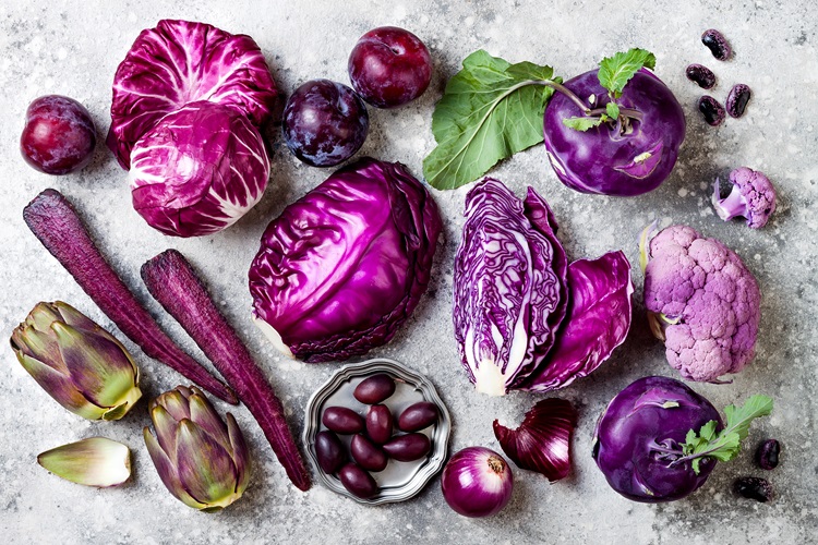 Purple vegetables over a grey background. Cabbage, radicchio salad, olives, kohlrabi, carrots, cauliflower, onions, artichoke, beans, potatoes, plums.