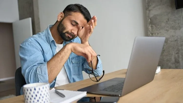 A tired man is working on his laptop and holding his head – behavioral symptoms of burnout syndrome that CBD oil can help with