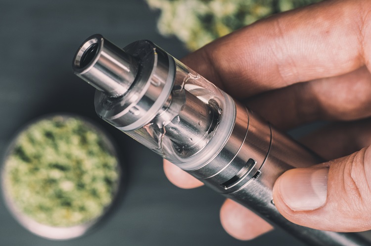 A hand holding a vaporizer with crushed CBD flowers in the background