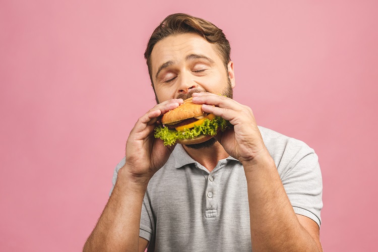 A man with closed eyes enjoys eating a burger against a pink background. He is wearing a gray polo shirt.