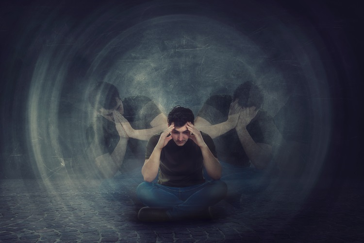 Man seated with head bowed, holding his head with his hands against a black background