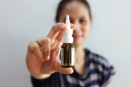 A person is holding a small bottle of nasal spray in front of them. The bottle has a white dispenser and is made of clear dark brown glass. The background is blurred, focusing on the bottle.